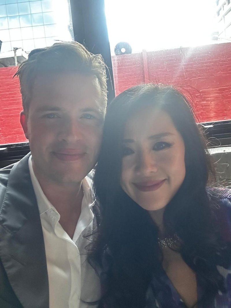 Close-up portrait of James Sackl and Alina Sackl in Sydney — candid shot, both smiling, with a red wall in the background.